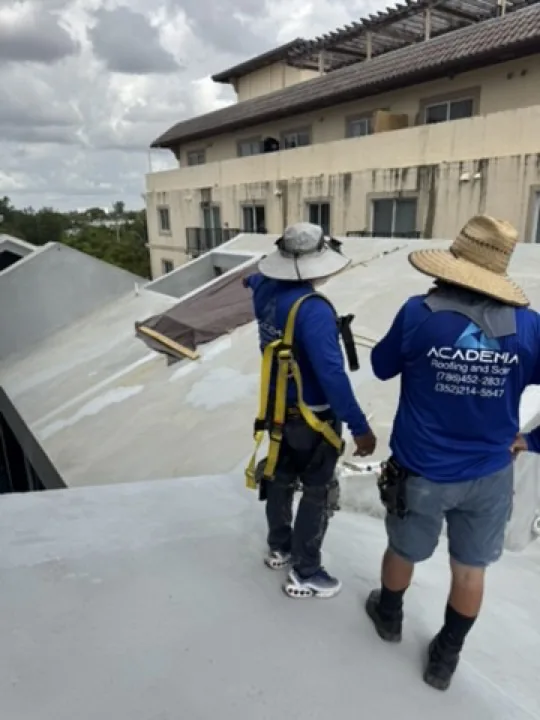 Two Academia-branded roofing workers wearing hats and fall-protection harnesses on a South Florida roof.